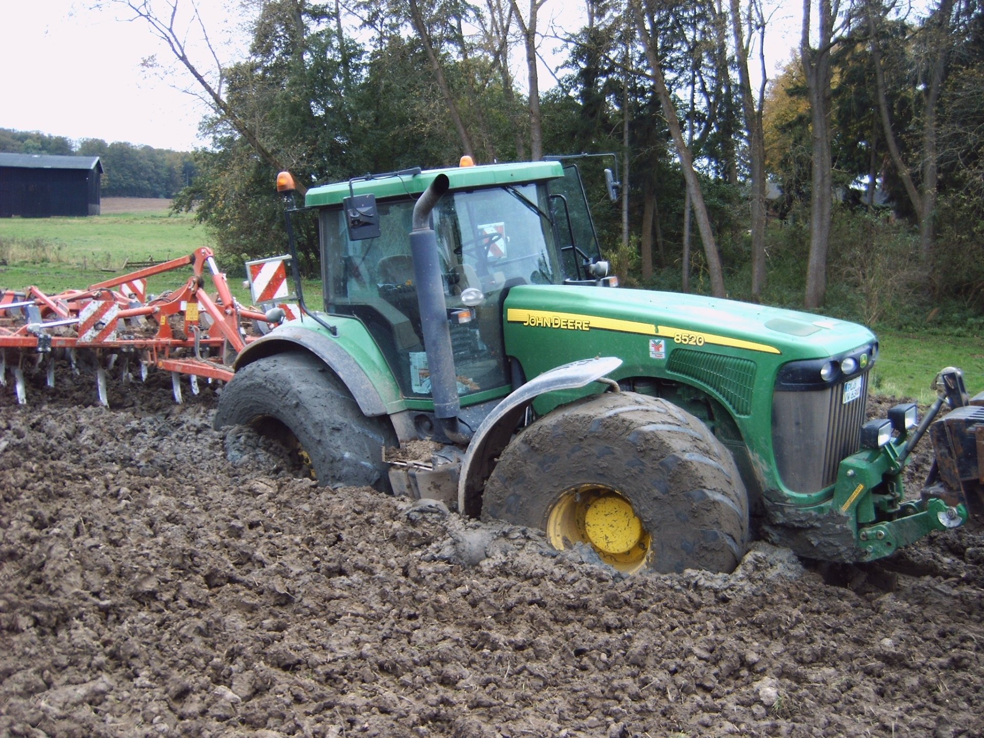 HD PC desktop wallpaper showing a John Deere vehicle partially stuck in muddy soil on a farm field surrounded by trees.