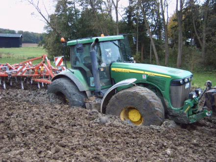 HD PC desktop wallpaper showing a John Deere vehicle partially stuck in muddy soil on a farm field surrounded by trees.