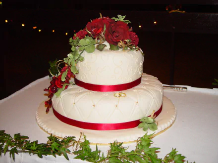 HD desktop wallpaper featuring a two-tiered wedding cake adorned with red roses, green leaves, and red ribbons, set against a dark background with soft lights.