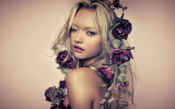 Portrait of blonde Australian model Gemma Ward with blue eyes, soft pink lipstick, and roses entwined in her hair, set against a neutral background.