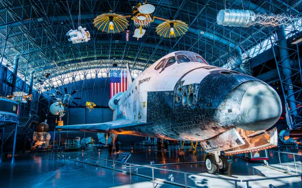 HD PC desktop wallpaper showing the Space Shuttle Discovery vehicle in a museum hangar, the orbiter lit beneath an arched glass roof with suspended spacecraft and an American flag.