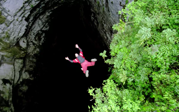 Base jumper in red freefall into a dark cave shaft beside lush green foliage, sports action captured as an HD PC desktop wallpaper and background.