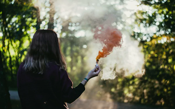 Rear-view of a person in a wood holding an orange smoke flare, misty light through trees — 4K Ultra HD photography desktop wallpaper/background.