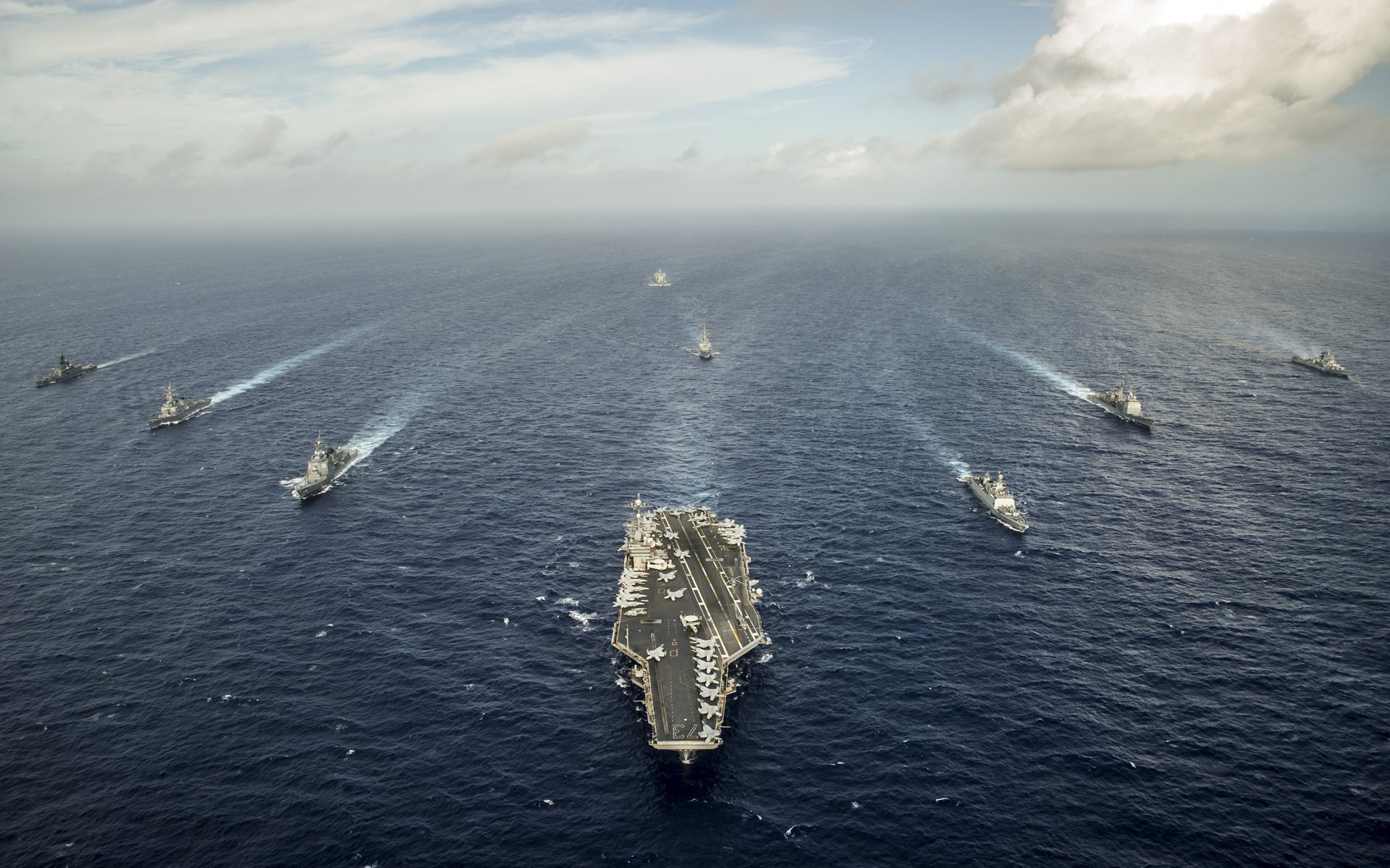 Aerial view of the USS George Washington (CVN-73) aircraft carrier leading a naval fleet across open ocean under a cloudy sky.