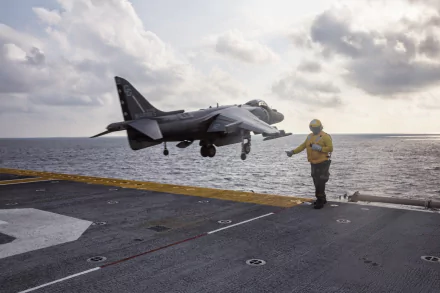 McDonnell Douglas AV-8B Harrier II jet fighter landing on USS Kearsarge with a Navy crew member directing, set against a cloudy sky and ocean backdrop.