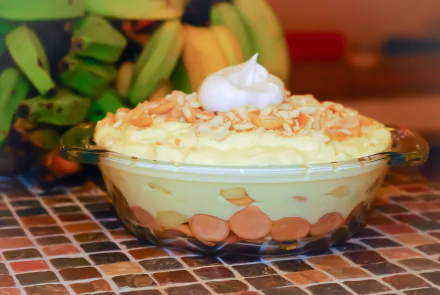 A close-up 4K Ultra HD wallpaper of creamy banana pudding topped with toasted coconut and whipped cream, with fresh bananas blurred in the background.