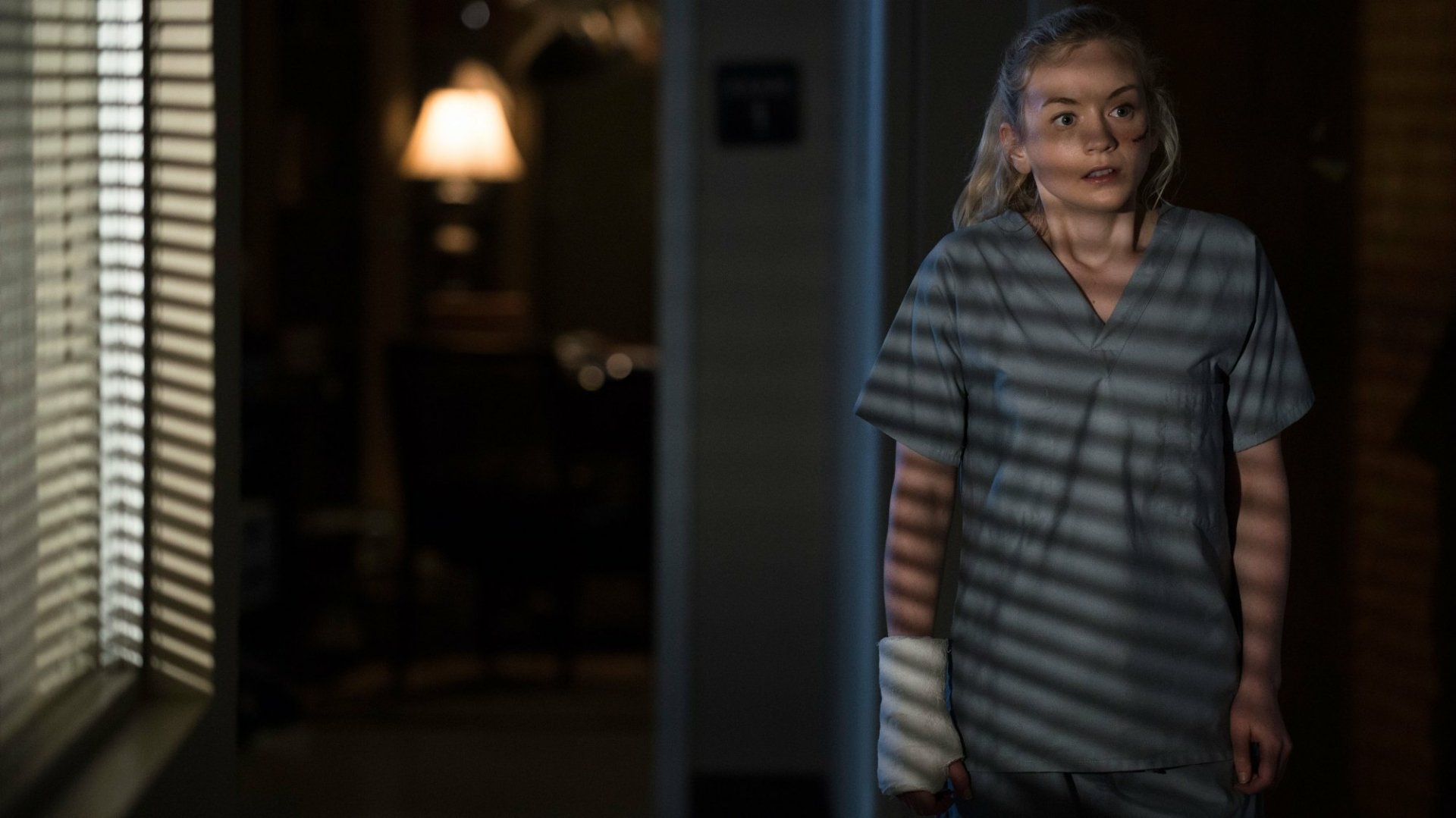 HD PC desktop wallpaper from the TV show The Walking Dead: a woman in scrubs with a bandaged arm stands in a dim room, blinds casting striped shadows across her.