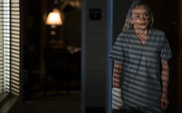 HD PC desktop wallpaper from the TV show The Walking Dead: a woman in scrubs with a bandaged arm stands in a dim room, blinds casting striped shadows across her.