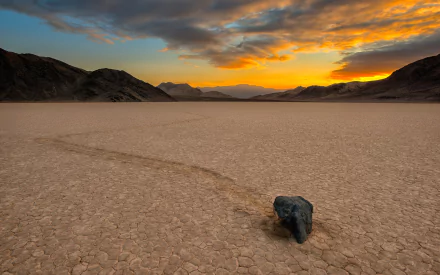 HD PC desktop wallpaper and background — Death Valley nature: a lone sliding rock leaving a trail across a cracked playa at sunset, distant mountains under dramatic clouds.