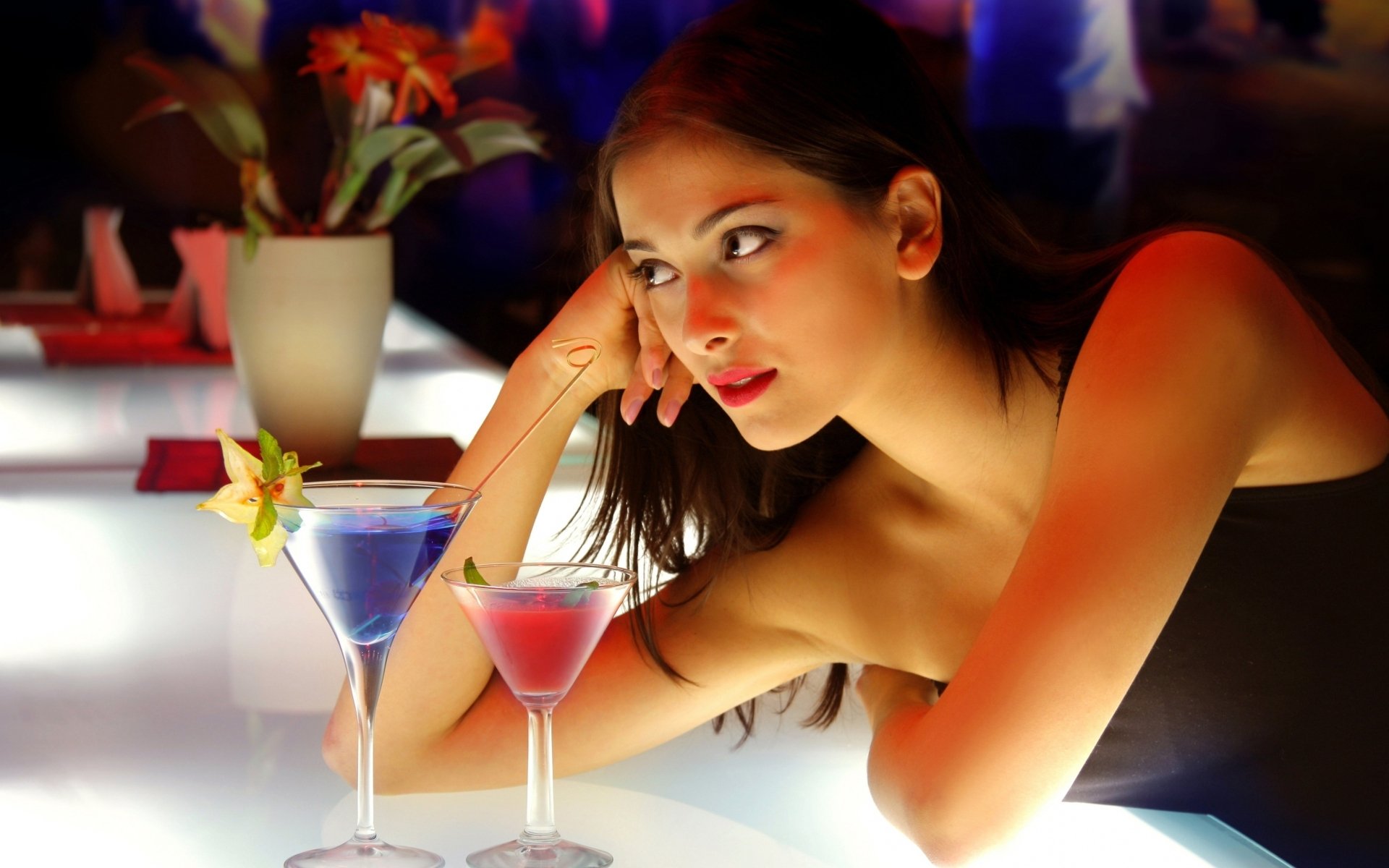 A stylish woman rests her chin on her hand, gazing thoughtfully at colorful cocktails on a vibrant bar, captured in an HD desktop wallpaper.