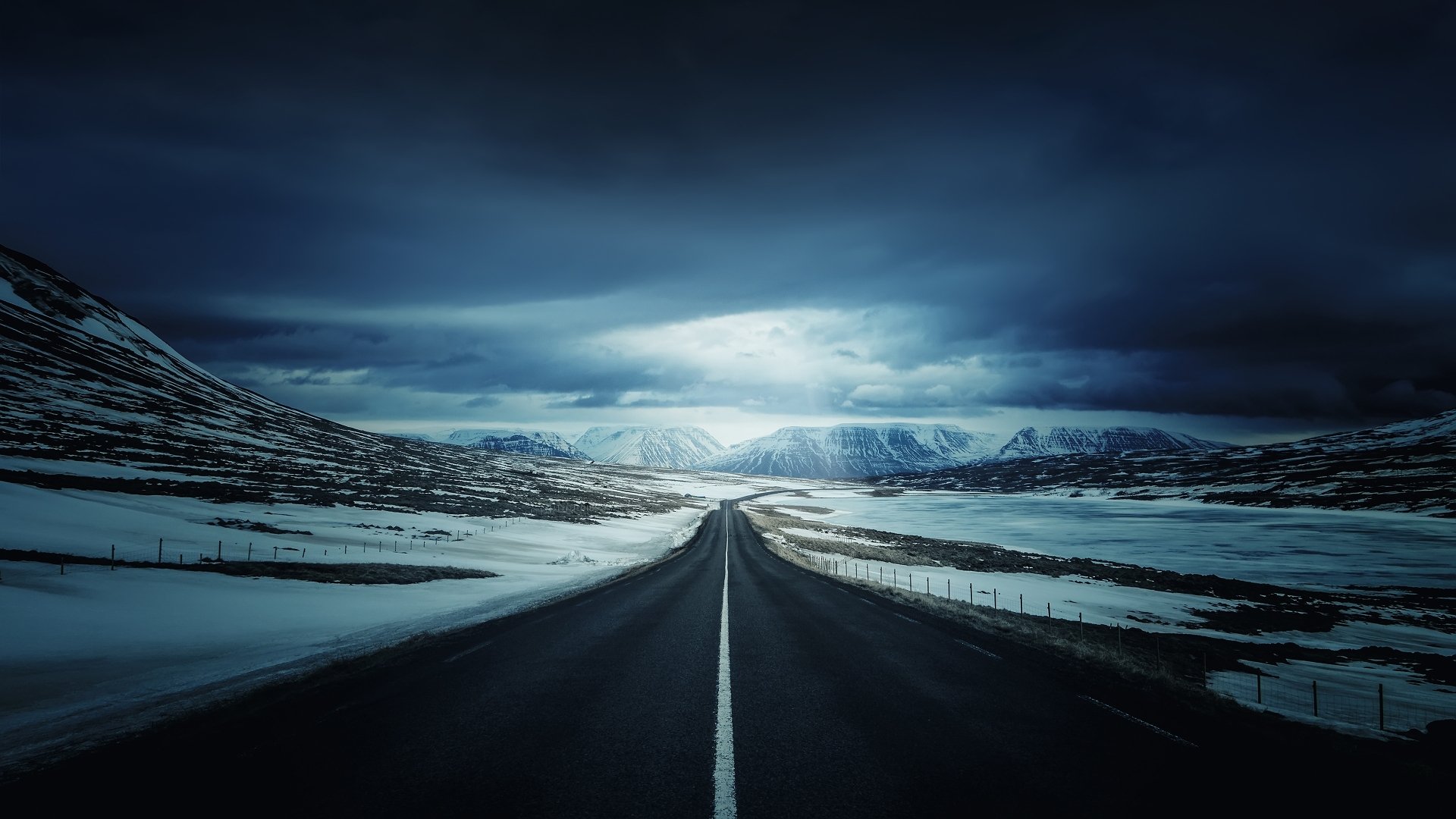 A high-definition night scene of a snow-covered mountain road in winter, framed by dark, dramatic skies. The road stretches into the distance, creating a sense of solitude and tranquility.