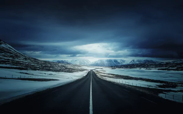 A high-definition night scene of a snow-covered mountain road in winter, framed by dark, dramatic skies. The road stretches into the distance, creating a sense of solitude and tranquility.