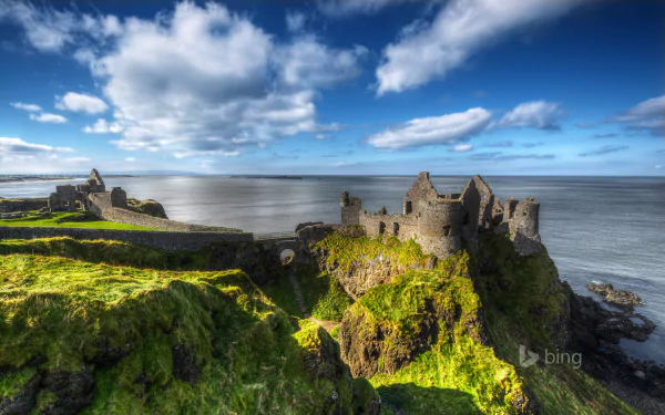 HD desktop wallpaper showcasing the man-made ruins of Dunluce Castle perched on a rugged coastline under a vibrant blue sky with scattered clouds.