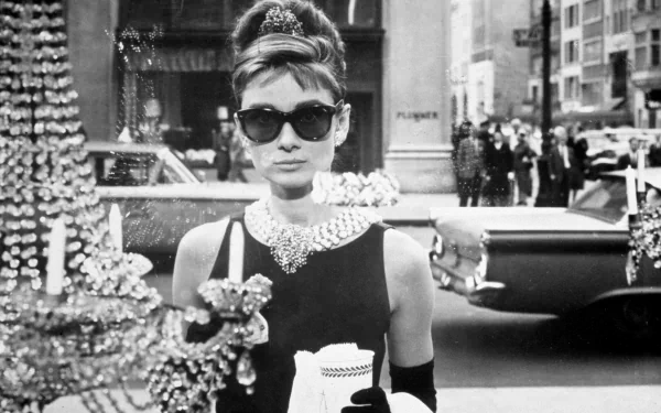 Black-and-white 4K PC desktop wallpaper of a glamorous mid‑century celebrity in a little black dress, pearls and oversized sunglasses, holding a long cigarette holder on a city sidewalk.