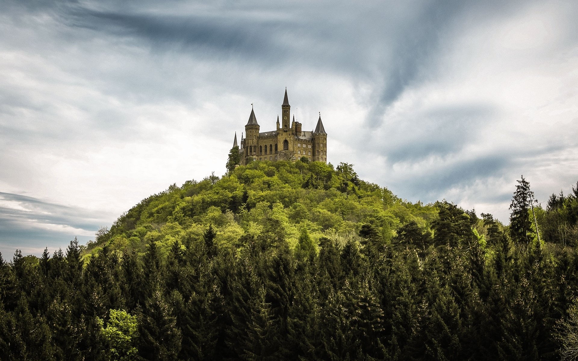 Hohenzollern Castle perched atop a lush green hill under a dramatic cloudy sky, captured in a high-definition desktop wallpaper.