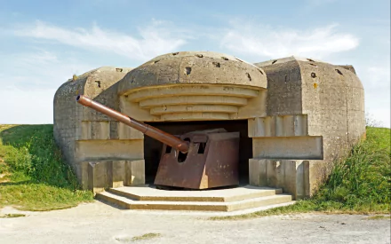HD PC desktop wallpaper: WWII-era concrete coastal military artillery bunker with a large gun protruding from its fortified opening, set on grassy terrain under a blue sky.