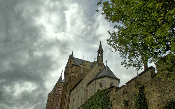 HD desktop wallpaper of Hohenzollern Castle with dramatic cloudy sky and lush greenery framing the historic man-made structure.