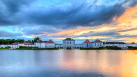 HD PC desktop wallpaper: Nymphenburg Palace, a man-made Baroque palace mirrored in calm water at sunset beneath dramatic clouds.