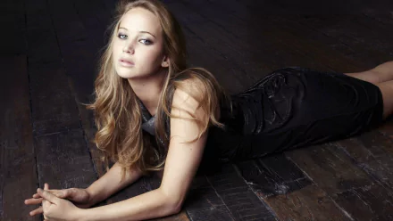 HD PC desktop wallpaper featuring celebrity Jennifer Lawrence lying on a dark wooden floor, gazing softly at the camera in an elegant black outfit.