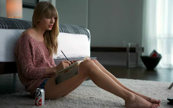 A HD desktop wallpaper featuring Taylor Swift sitting on the floor, writing in a notebook, with a cozy atmosphere and a soda can beside her.