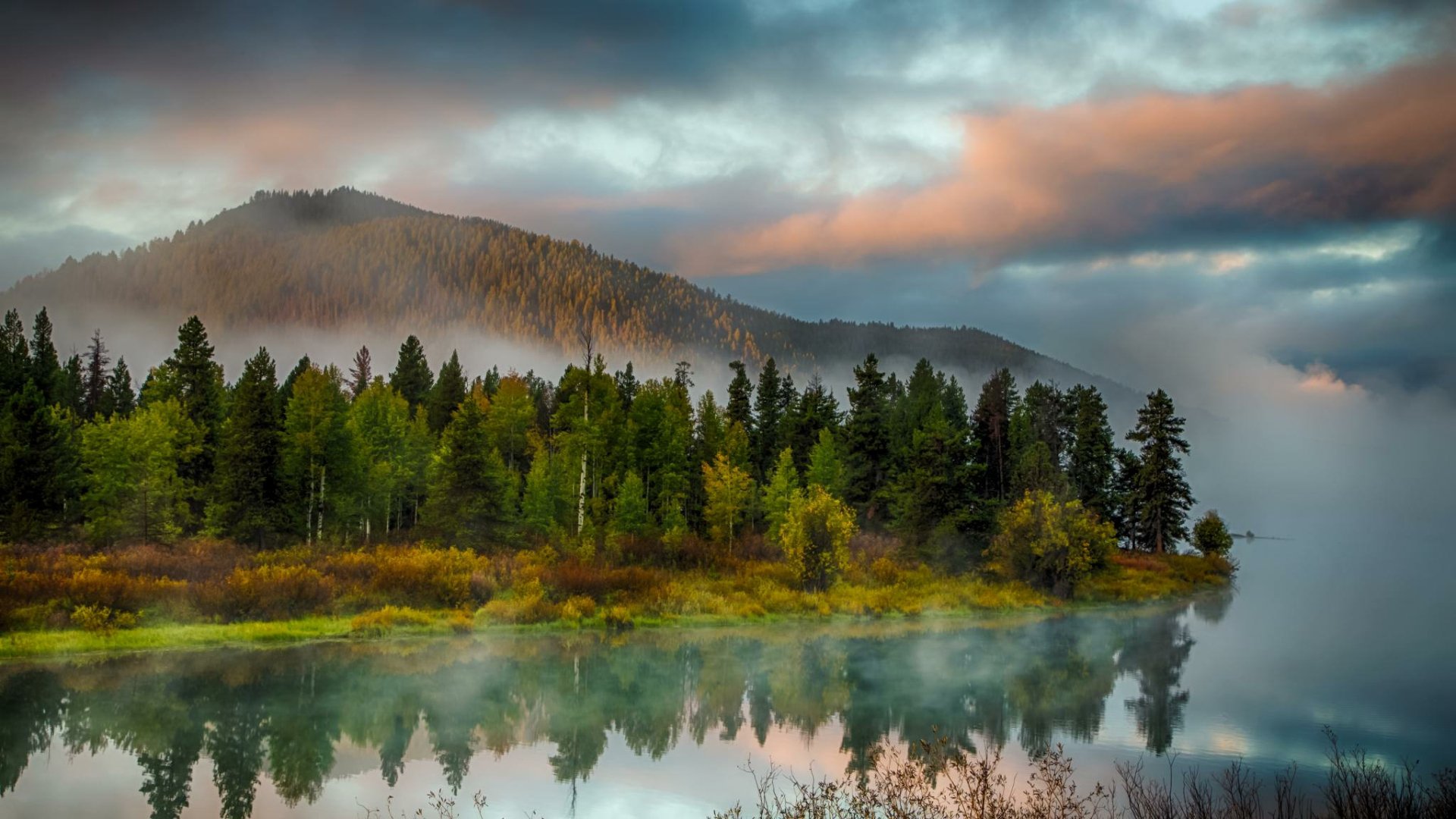 HD PC desktop wallpaper showing a serene landscape with a misty lake, dense evergreen trees, and a mountain under a moody, colorful sky at dawn.