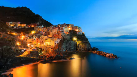 A stunning HD wallpaper of Manarola, showcasing its colorful buildings illuminated at twilight, perched on a rugged coastline with calm waters reflecting the vibrant lights.