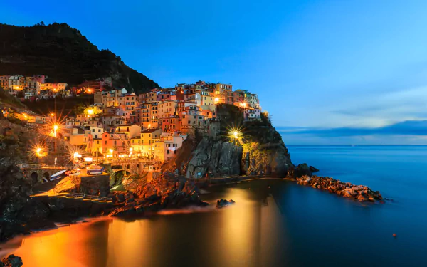 A stunning HD wallpaper of Manarola, showcasing its colorful buildings illuminated at twilight, perched on a rugged coastline with calm waters reflecting the vibrant lights.