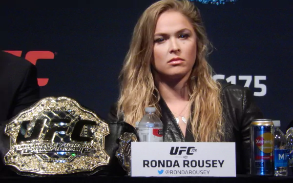 Ronda Rousey sits at a press conference, displaying her championship belt. This HD wallpaper highlights her intense focus and determination in the world of sports.