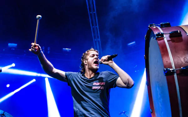 A dynamic performance by Imagine Dragons, an American alternative band, capturing the energy of live music with a vocalist and a large drum on stage, illuminated by vibrant lights.