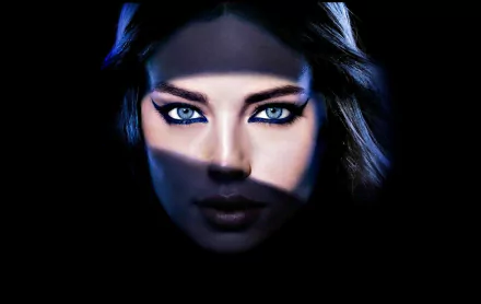 A striking HD wallpaper featuring the captivating face of a celebrity, Emily Didonato, with intense blue eyes partially illuminated in shadow.