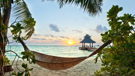 A serene tropical beach at sunset, featuring a hammock strung between palm trees and a gazeboon the shore, with the colorful sea and sky creating a stunning backdrop. 