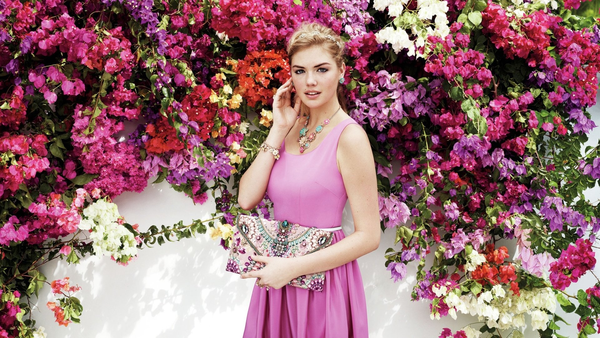 Celebrity Kate Upton poses in a vibrant pink dress holding a stylish bag, surrounded by colorful blossoming flowers in this HD PC desktop wallpaper.