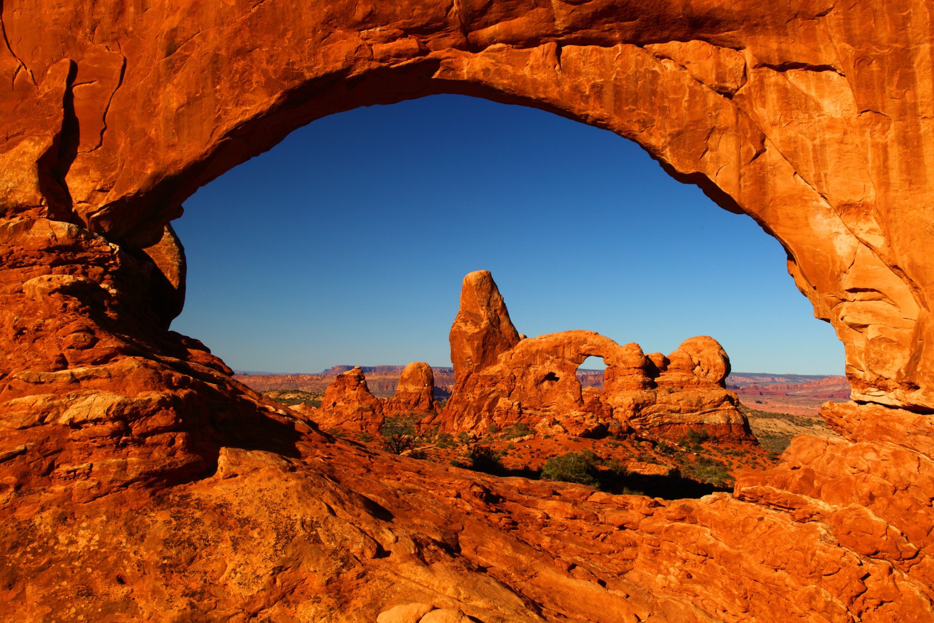 HD PC desktop wallpaper: sunlit red sandstone arch framing a distant spire and arches over a desert canyon under a clear blue sky.