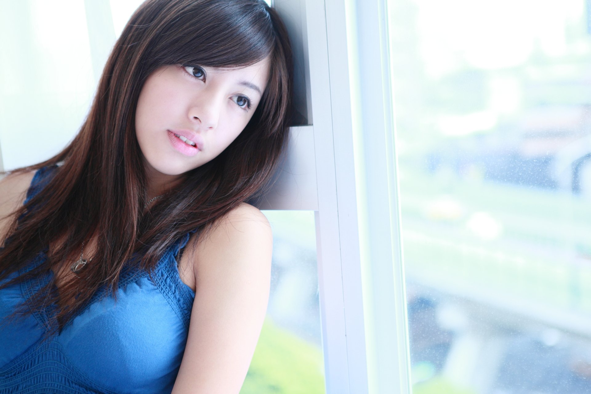 HD Wallpaper featuring Gū Guǎnyǔn, an oriental Taiwanese woman, gazing thoughtfully outside a window. She has long, straight black hair and is wearing a blue top.