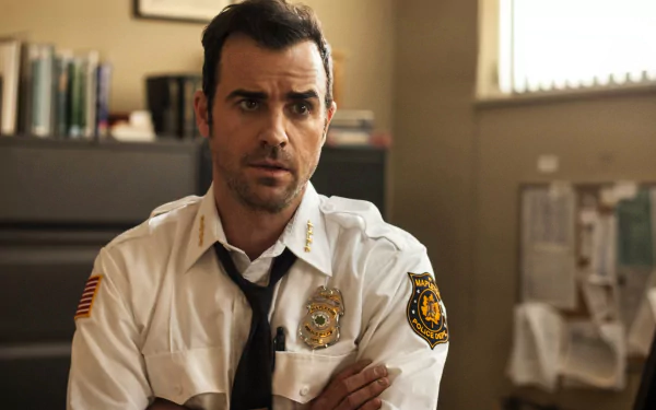 HD PC desktop wallpaper from TV show The Leftovers: a tense male sheriff in uniform sits with arms crossed in a cluttered office, books and papers visible behind him.