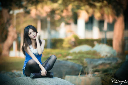 A woman named Julie Chang, of Taiwanese descent, poses gracefully on a rock, surrounded by a blurred, serene outdoor setting, capturing a moment in soft focus.