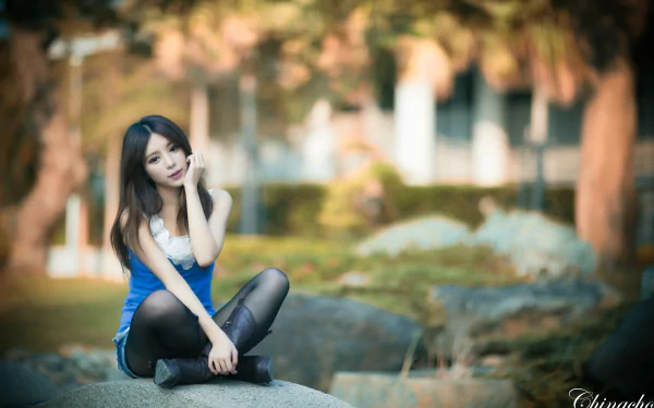 A woman named Julie Chang, of Taiwanese descent, poses gracefully on a rock, surrounded by a blurred, serene outdoor setting, capturing a moment in soft focus.