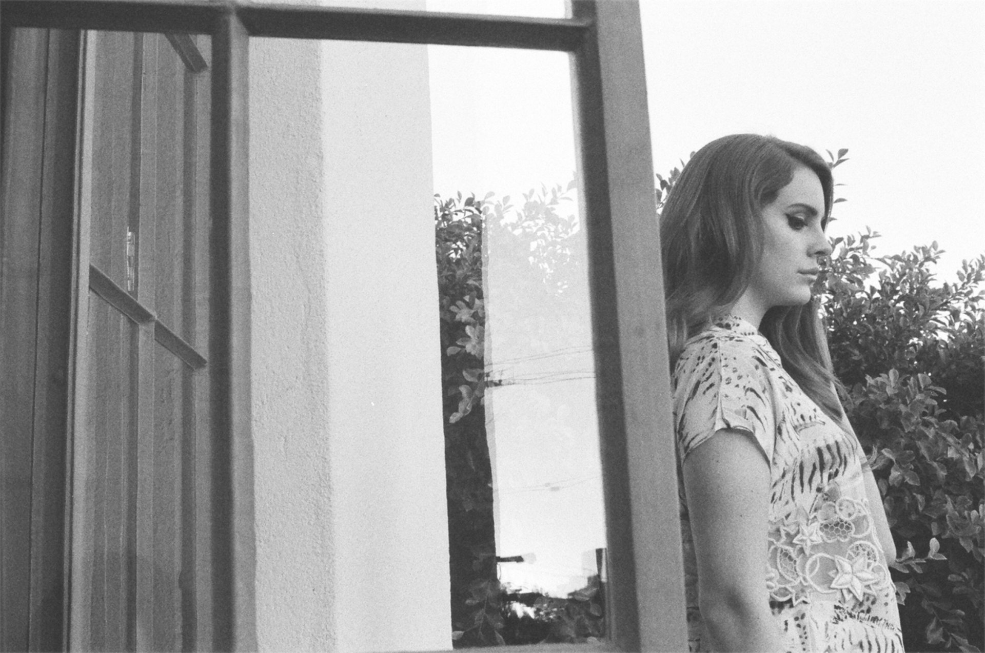 Black and white HD PC desktop wallpaper featuring Lana Del Rey standing thoughtfully by a window, with natural outdoor foliage in the background.