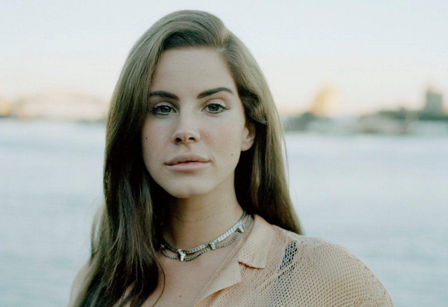 HD desktop wallpaper featuring a close-up portrait of Lana Del Rey against a soft, blurred outdoor background, evoking a serene and musical vibe.
