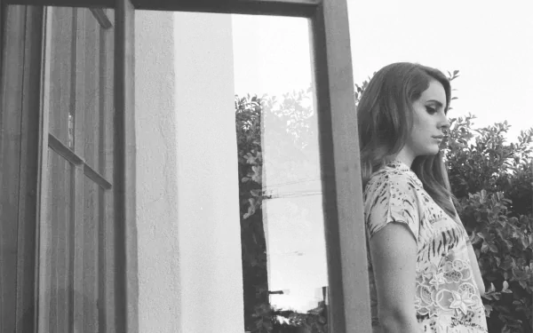 Black and white HD PC desktop wallpaper featuring Lana Del Rey standing thoughtfully by a window, with natural outdoor foliage in the background.