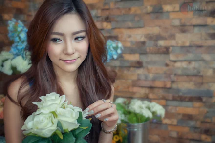 A smiling Thai woman with long hair holds a bouquet of flowers, set against a rustic wooden backdrop filled with blooms. An elegant HD wallpaper for desktop backgrounds.