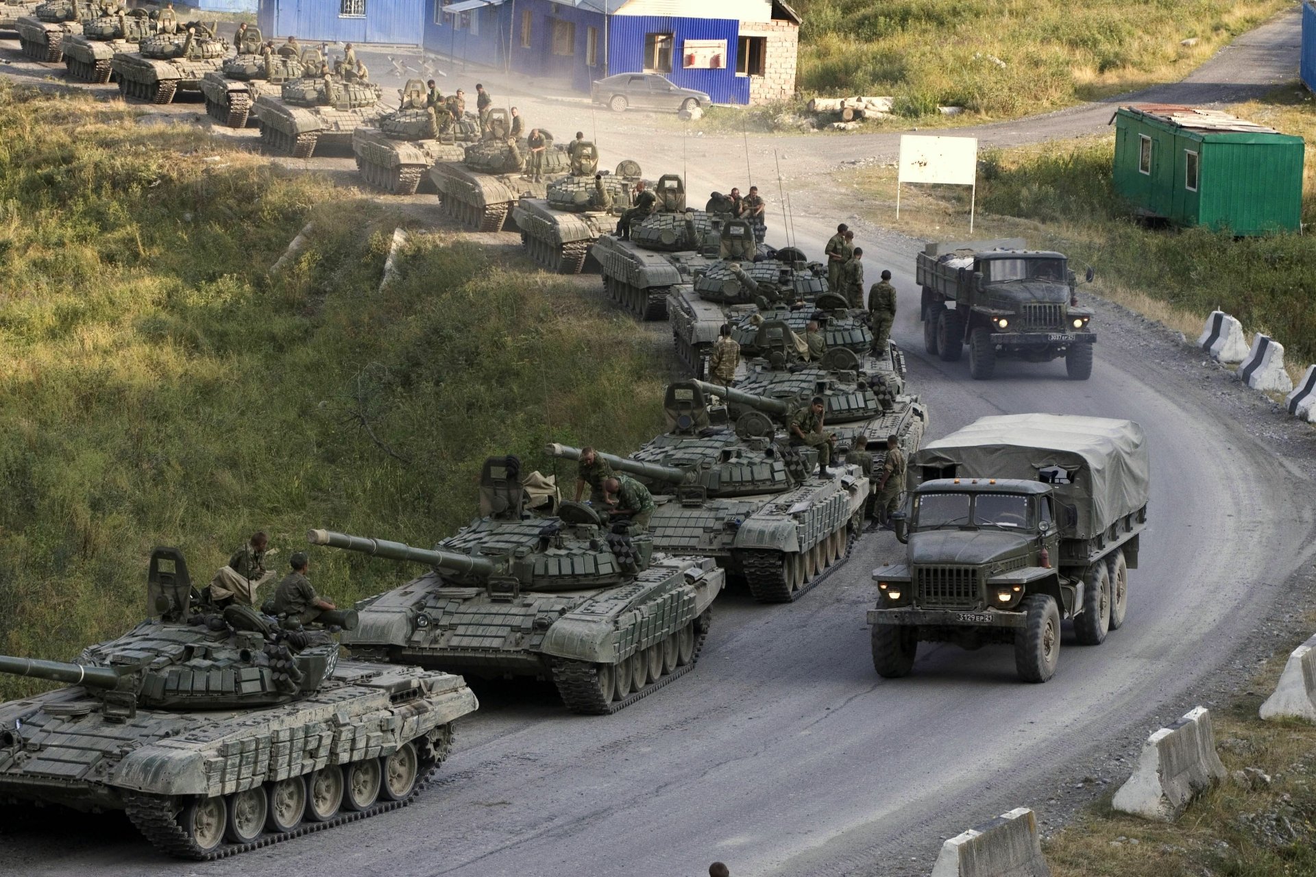 A line of military tanks drives along a road, with soldiers on board and an army truck nearby, set against a backdrop of grass and military structures. High-definition wallpaper.