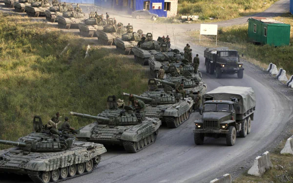 A line of military tanks drives along a road, with soldiers on board and an army truck nearby, set against a backdrop of grass and military structures. High-definition wallpaper.