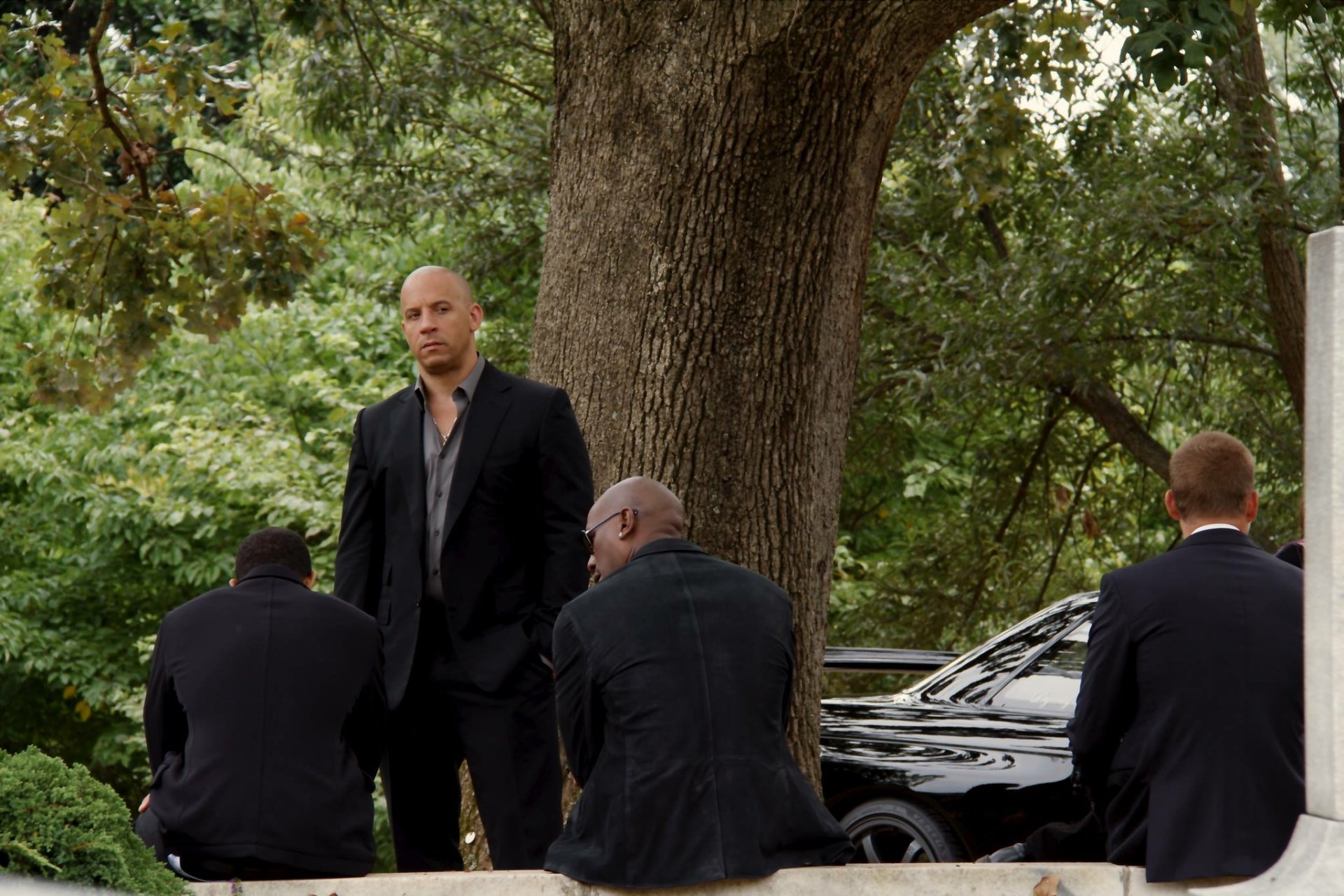 Dominic Toretto, portrayed by Vin Diesel, stands confidently amidst a lush background with allies in suits, from the action-packed movie Furious 7. HD desktop wallpaper.