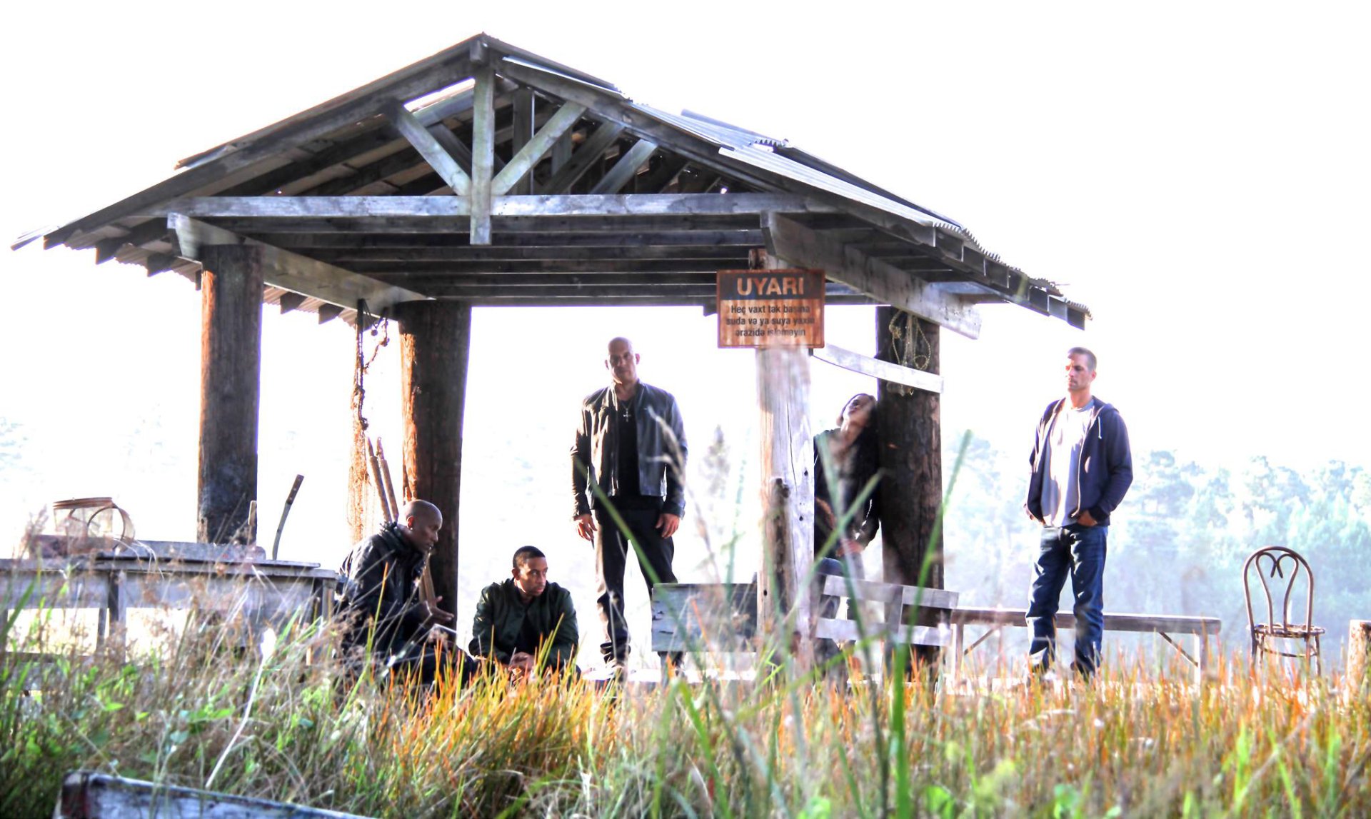 A scene from Furious 7 featuring characters Letty Ortiz, Tej, Roman Pearce, and Brian O'Conner gathered at a rustic pavilion, capturing a moment of camaraderie and tension.
