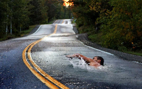 HD desktop wallpaper featuring a manipulated image of a man swimming in a water-covered section of a winding road surrounded by trees at sunset.