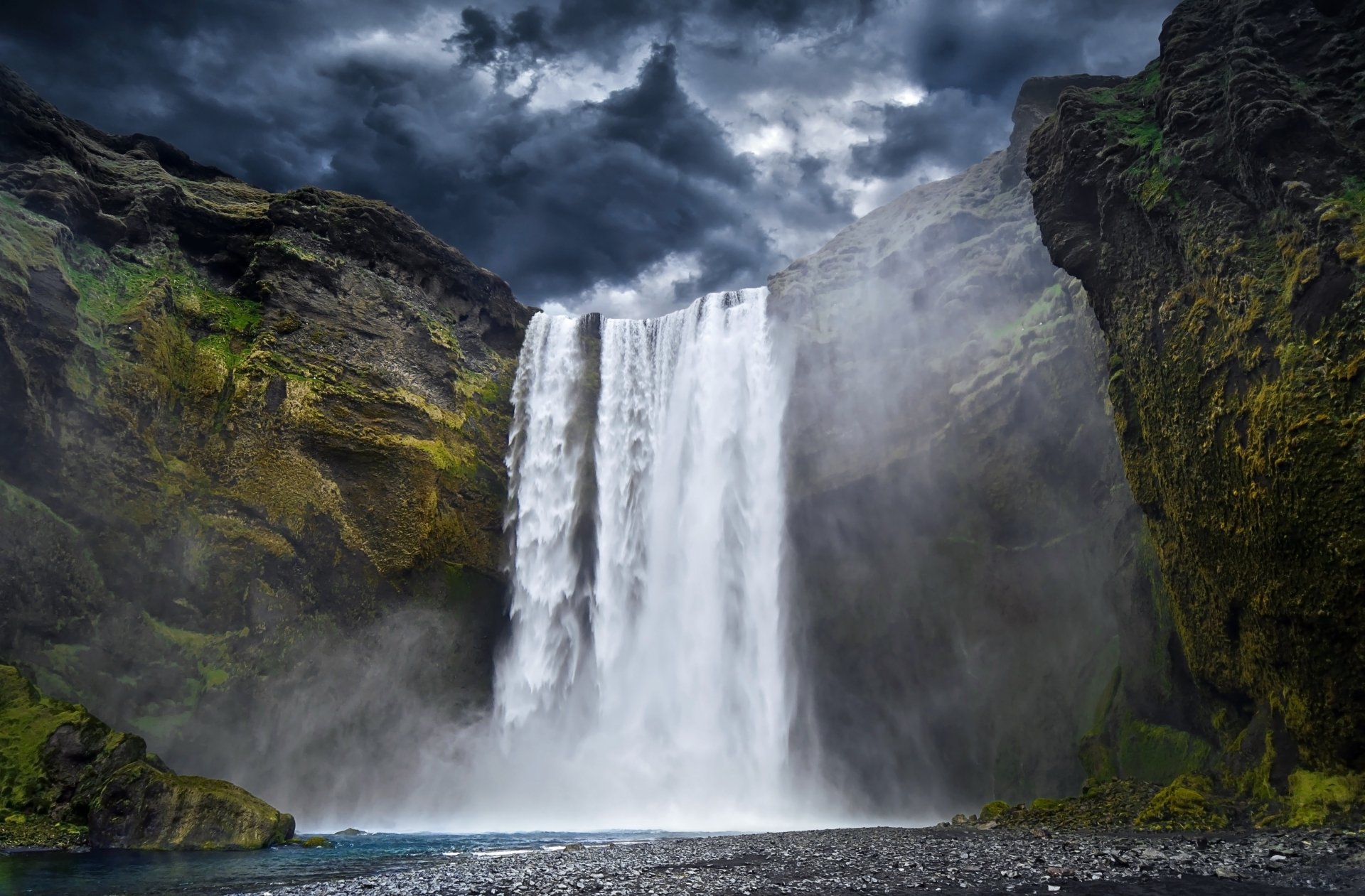 Skógafoss waterfall in Iceland cascades powerfully under dramatic cloud-filled skies, surrounded by lush moss-covered cliffs, captured in stunning 4K Ultra HD quality.