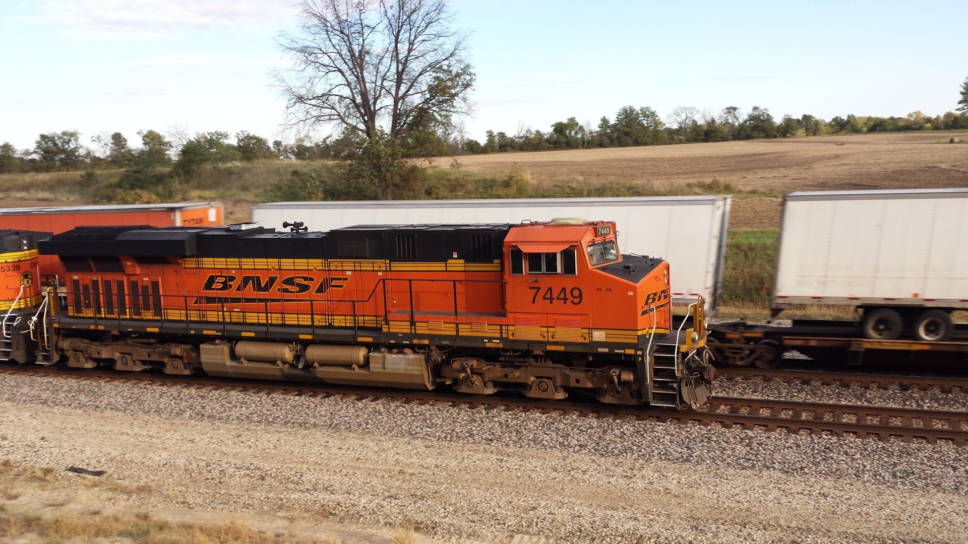 A vibrant orange BNSF train locomotive moves along tracks beside parked freight containers, set against a backdrop of open fields and trees, captured in stunning 4K Ultra HD quality.