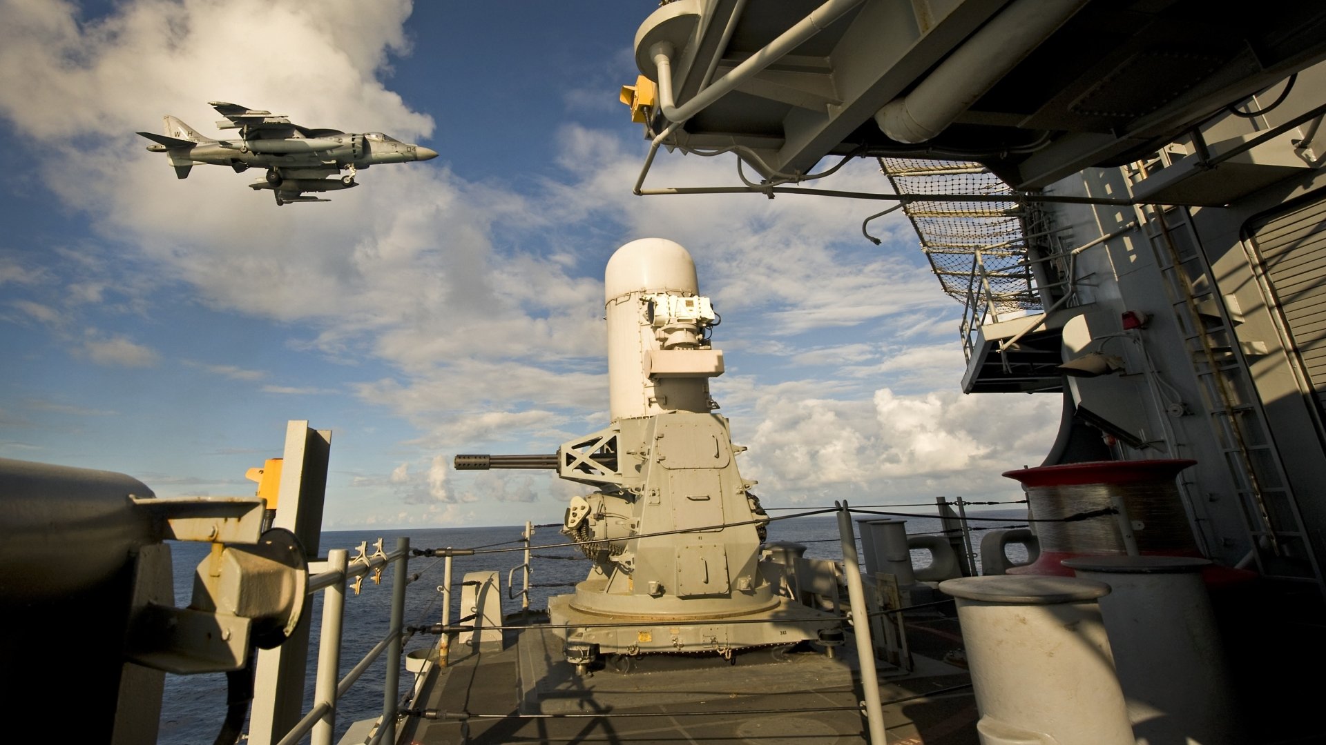 HD desktop wallpaper featuring a McDonnell Douglas AV-8B Harrier II military jet flying over a naval ship deck under a partly cloudy sky.