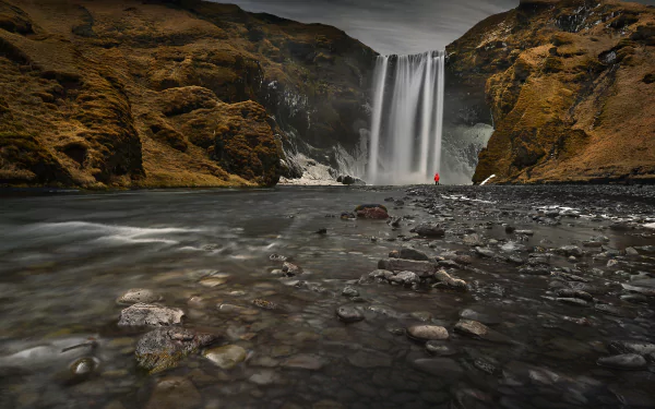 HD desktop wallpaper of Skógafoss waterfall in Iceland during spring, showcasing flowing water and rugged nature with a person in red near the waterfall.